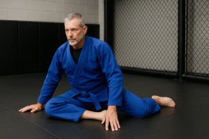 BJJ Hip Mobility Stretch for Grapplers Over 40 OLder BJJ practitioner in a blue gi preforming a seated hip mobilitystrechon the mats