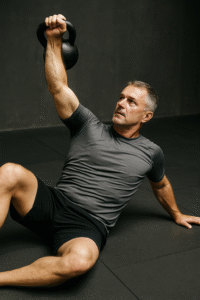 Older male BJJ athlete performing a kettlebell armbar drill in no-gi, wearing grey rashguard and black shorts.